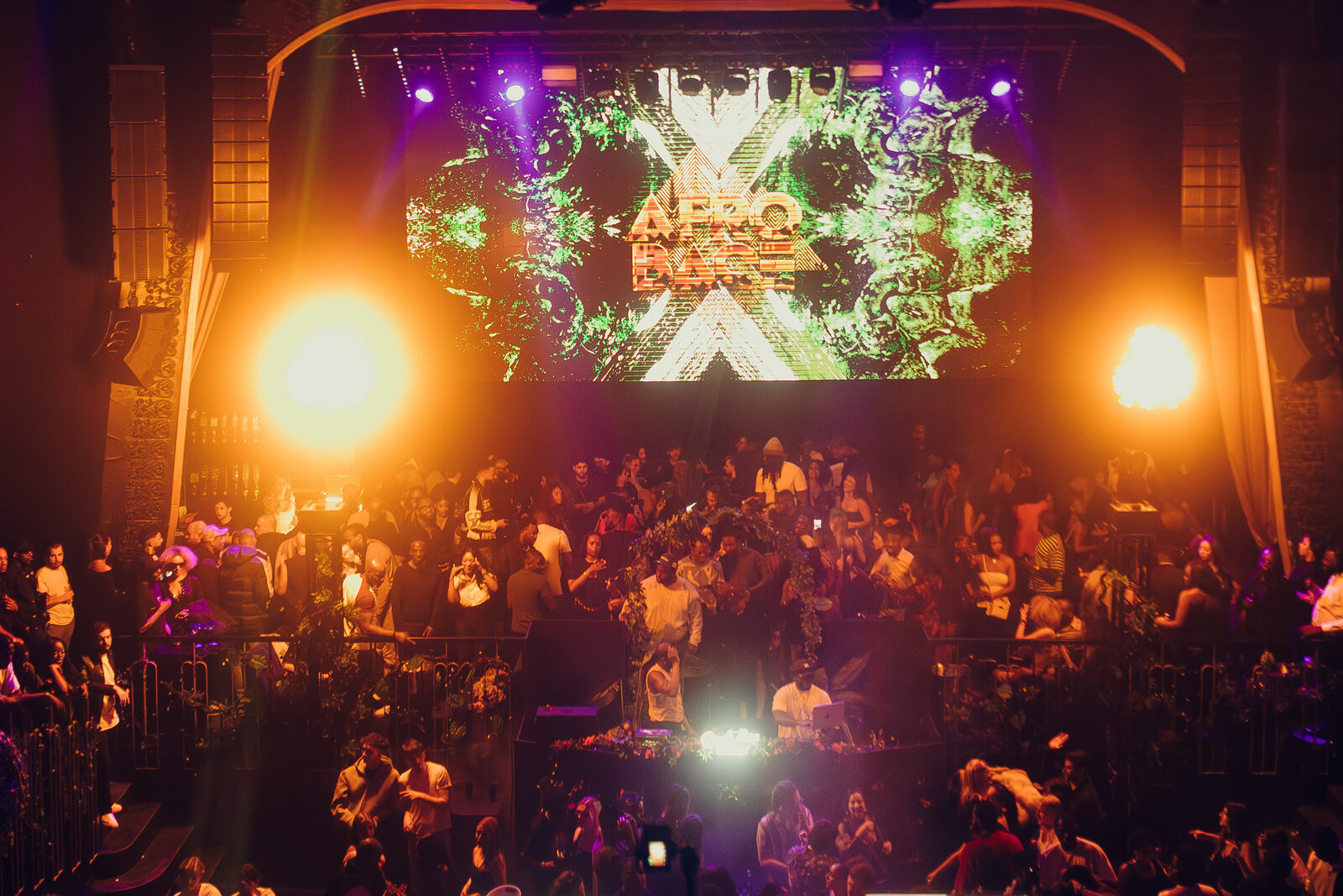 A packed crowd dancing in a nightclub beneath a giant illuminated 'Afrobase' screen, with two DJs performing under warm stage lighting.