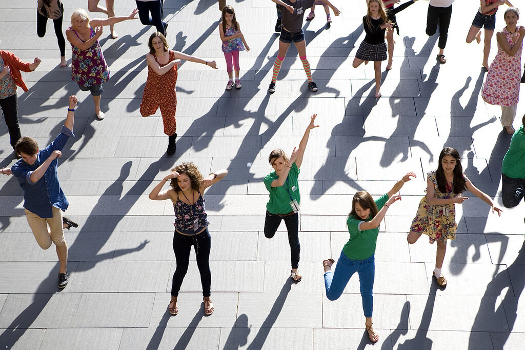 An overhead view of a large group of people of all ages performing a synchronised public dance routine on a sunlit square.