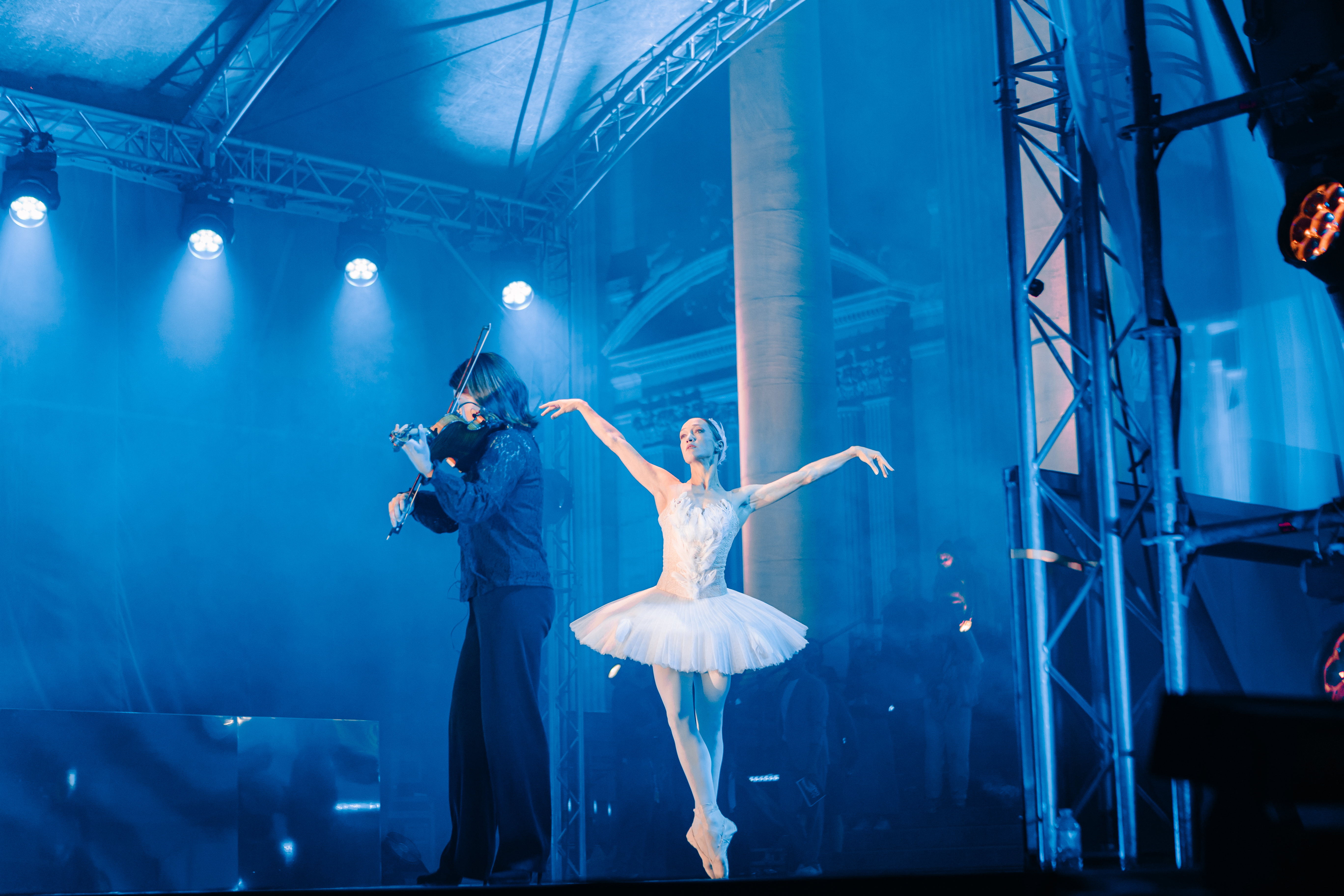 A ballerina in a white tutu dancing en pointe alongside a violinist on an open-air stage in Brussels, under blue stage lighting and a metal truss structure