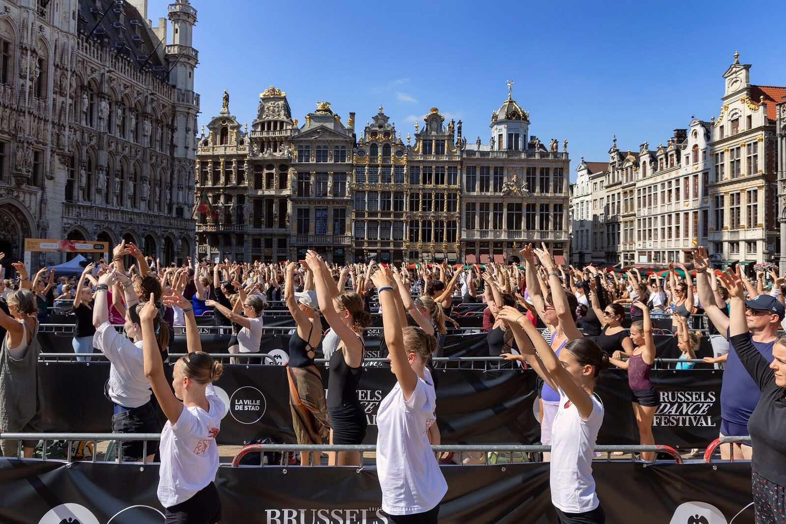 A mass ballet barre class filling the Grand-Place in Brussels, dancers with arms raised in front of the square's historic guild houses, with Brussels Dance Festival banners visible.