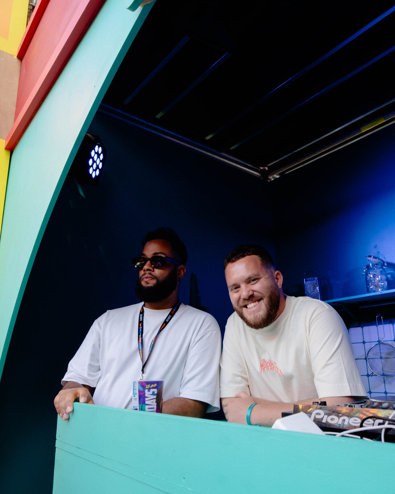 Two DJs smiling behind a Pioneer DJ set-up in a colourful venue booth.