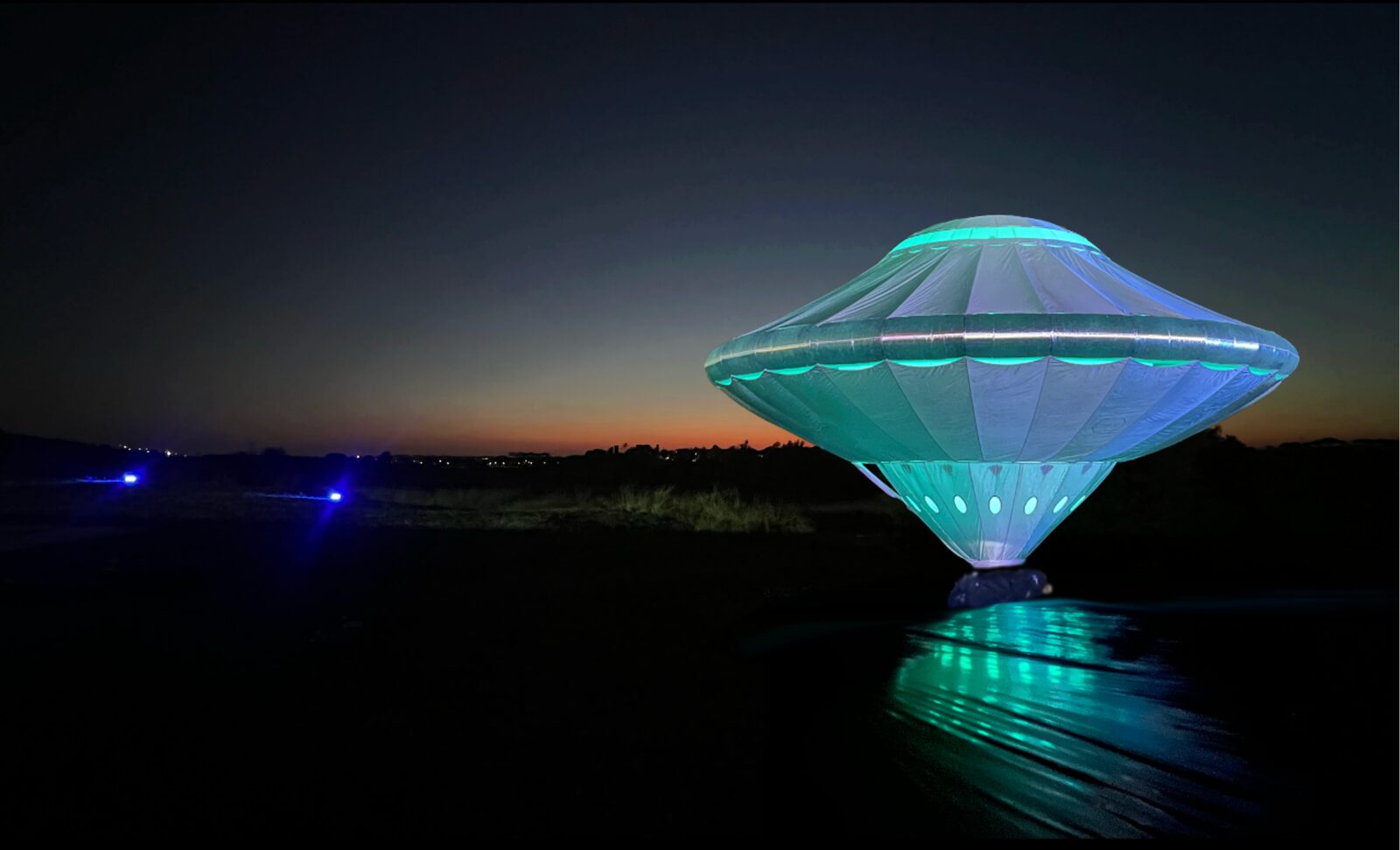 A giant illuminated flying-saucer sculpture glowing turquoise and white against a dusk sky, its light reflected on the ground below