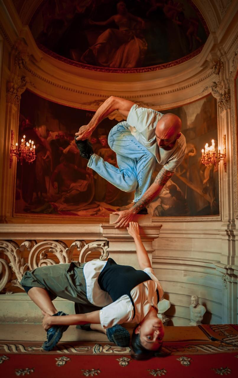 Two breakdancers mid-move in a grand, ornately decorated historic hall at the Hôtel de Ville, one balancing on a stone ledge, the other in a low floor pose
