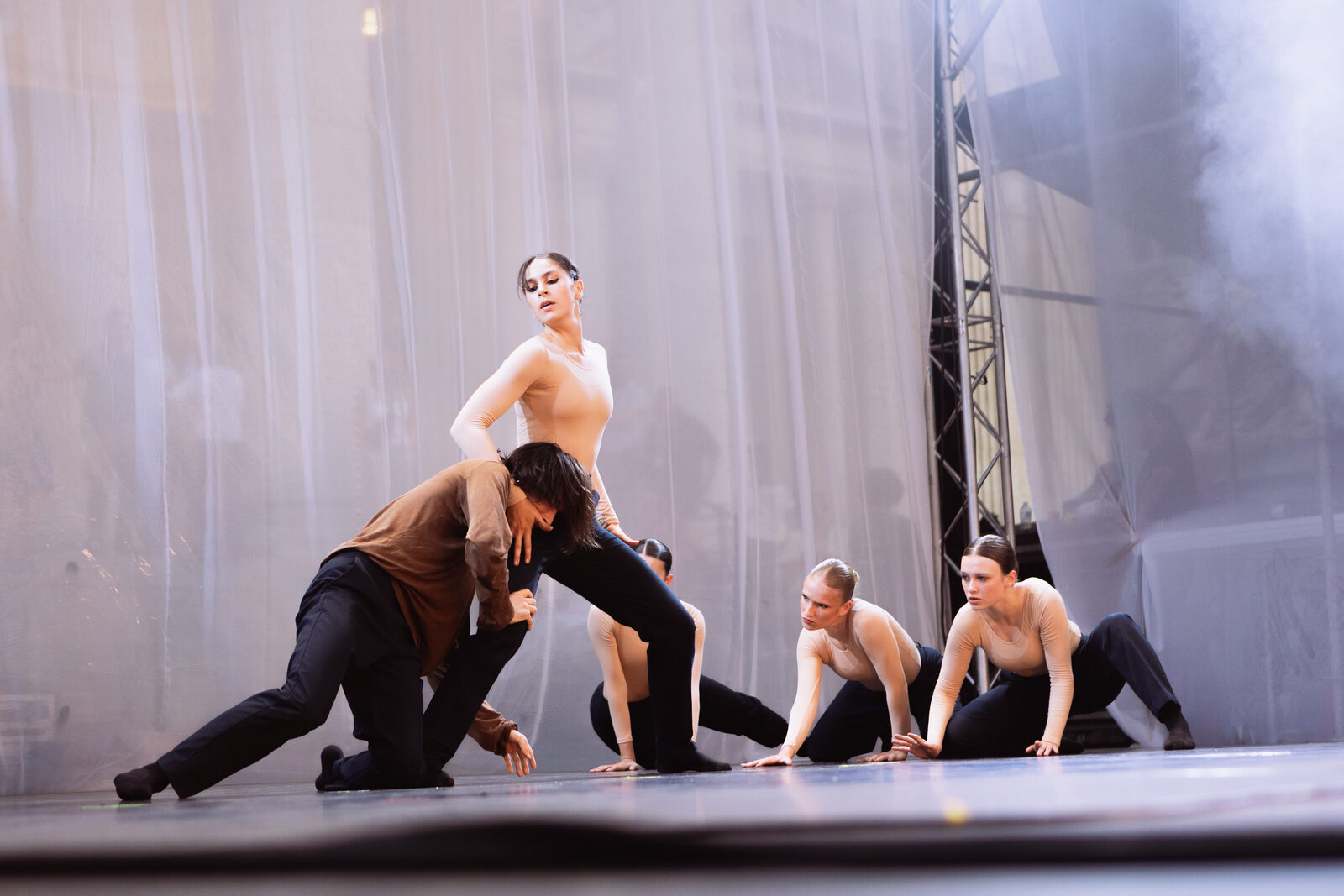 A group of dancers crouched low on stage in a synchronised pose, in front of a soft sheer curtain backdrop under a metal truss structure