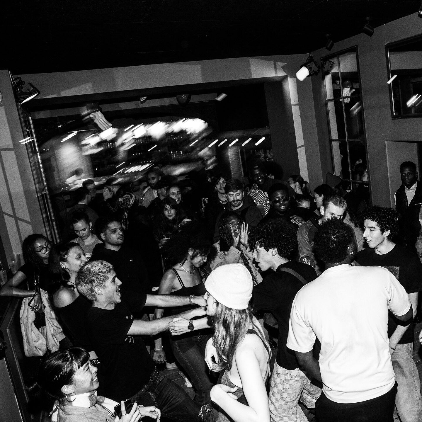 A crowded, dimly lit dancefloor packed with people dancing and laughing together, in black and white.