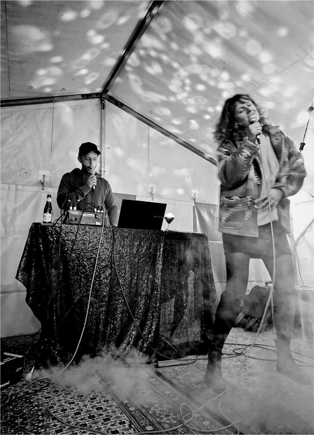 Two performers singing into microphones under a party tent filled with fog and disco-ball light.
