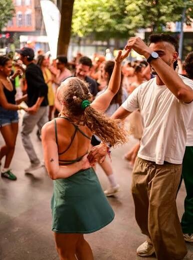 A couple salsa dancing hand-in-hand in a crowded outdoor square, the woman mid-spin